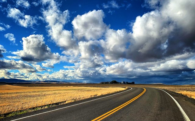 Open highway curving through golden prairie fields under a big cloudy sky