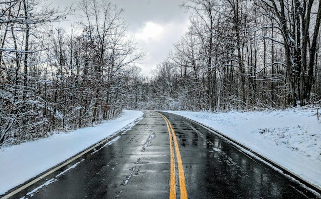 Wet two-lane road with a double yellow line between snowy banks and bare trees