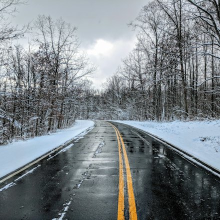 Wet two-lane road with a double yellow line between snowy banks and bare trees