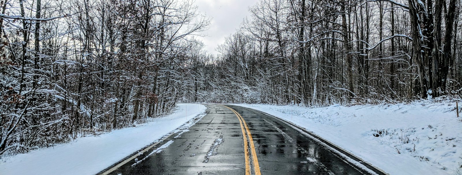 Wet two-lane road with a double yellow line between snowy banks and bare trees